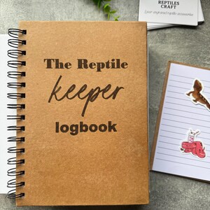 May include: A brown spiral-bound notebook with the text "The Reptile Keeper Logbook". Beside it are business cards with a reptile logo and a small notepad with reptile stickers. The notebook is on a grey surface.