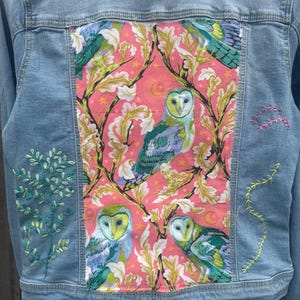 May include: Light blue denim jacket with a vibrant pink panel featuring owl illustrations and floral patterns. The jacket has embroidered details on the sleeves and back, showcasing a unique, artistic design.