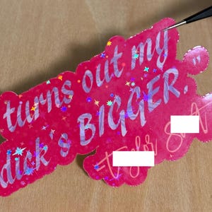 May include: A pink sticker with the text "turns out my dick's BIGGER." The sticker has a holographic effect with star-shaped accents. The sticker is being held by a pair of tweezers.