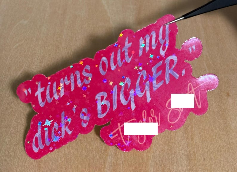 May include: A pink sticker with the text "turns out my dick's BIGGER." The sticker has a holographic effect with star-shaped accents. The sticker is being held by a pair of tweezers.