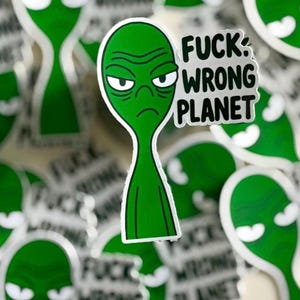 May include: A collection of green alien stickers with a grumpy expression. One sticker features the text "FUCK: WRONG PLANET" in black and white. The stickers have a white border and are scattered across the image.