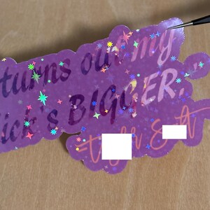 May include: A purple sticker with the text "turns out my dick's BIGGER" in a playful font. The sticker has a scalloped edge and is decorated with colorful star-shaped accents. A pair of tweezers is touching the sticker.