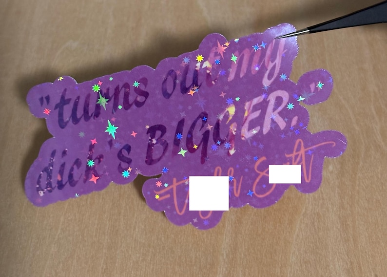 May include: A purple sticker with the text "turns out my dick's BIGGER" in a playful font. The sticker has a scalloped edge and is decorated with colorful star-shaped accents. A pair of tweezers is touching the sticker.