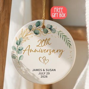 Puede incluir: Un adorno redondo de cerámica blanca con un diseño de hojas de eucalipto doradas y verdes. El texto dice 20th Anniversary, James & Susan y July 29 2026, con dos corazones dorados. Incluye un cordón dorado para colgar y una etiqueta roja.