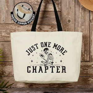 Puede incluir: Un bolso tote de lona color crema con asas negras y un estampado gráfico negro de un esqueleto leyendo un libro. El texto dice "JUST ONE MORE CHAPTER" con detalles de estrellas. Un recuadro circular muestra un bolsillo interior con cremallera.