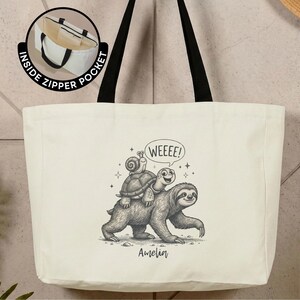 May include: A cream-colored tote bag with black handles featuring a black and white sketch of a sloth carrying a turtle and a snail. The text WEEEE! appears in a speech bubble above the animals, and the name Amelia is printed below the illustration.