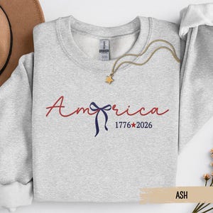 May include: Ash-colored sweatshirt with the word "America" in red script and a blue bow graphic. The text "1776*2026" is below the word "America". A gold necklace with a star pendant is on the sweatshirt.