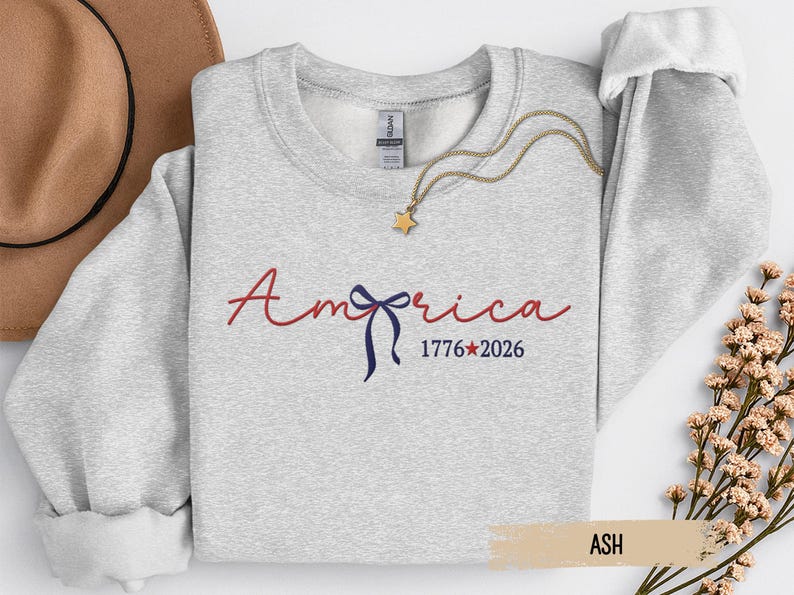 May include: Ash-colored sweatshirt with the word "America" in red script and a blue bow graphic. The text "1776*2026" is below the word "America". A gold necklace with a star pendant is on the sweatshirt.