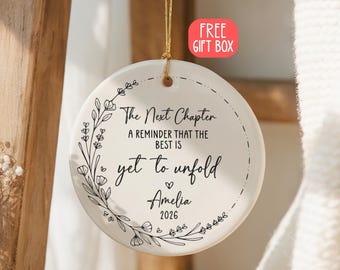May include: A round white ceramic ornament hanging by a gold string. It features a black floral wreath design and the text: The Next Chapter, A REMINDER THAT THE BEST IS yet to unfold, Amelia, 2026. The ornament has a smooth, glossy finish.