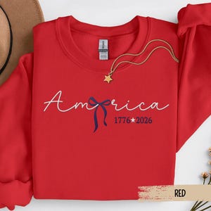 May include: A red sweatshirt with the word "America" in white script, a blue bow, and the dates "1776*2026". A gold star pendant hangs from a gold chain. The sweatshirt is folded with a brown hat visible.