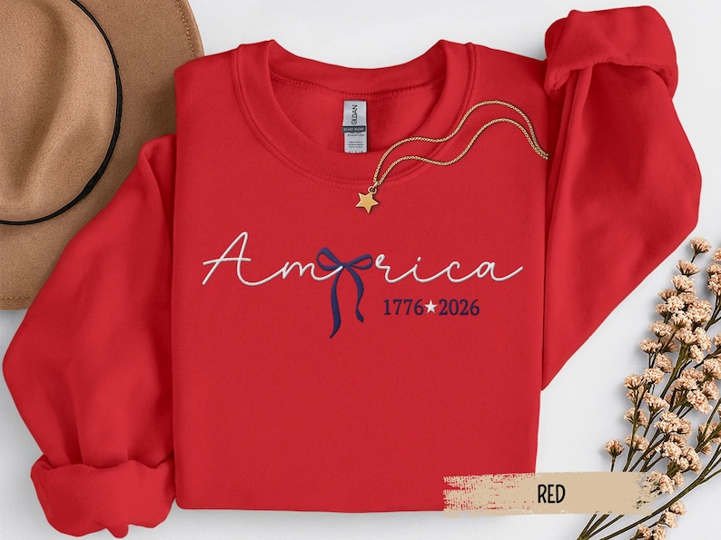 May include: A red sweatshirt with the word "America" in white script, a blue bow, and the dates "1776*2026". A gold star pendant hangs from a gold chain. The sweatshirt is folded with a brown hat visible.