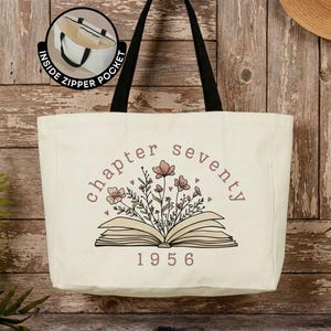 May include: A cream-colored canvas tote bag with black shoulder straps. The front features a graphic of an open book with wildflowers emerging from the pages, the text "chapter seventy" arched above, and "1956" below. An inset shows an inside zipper pocket.