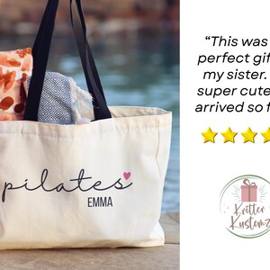 Pu&ograve; includere: Una borsa tote in tela bianca con manici neri che presenta il testo nero pilates ed EMMA con un piccolo cuore rosa. All'interno della borsa ci sono un panno con motivo arancione e bianco e un panno con motivo geometrico blu e bianco.