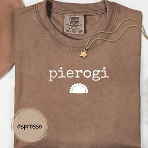 Könnte beinhalten: Braunes T-Shirt mit dem weißen Wort „pierogi“ und einer Pierogi-Grafik. Eine goldene Stern-Anhänger-Halskette und ein brauner Hut sind ebenfalls zu sehen. Das Shirt ist mit „Comfort Colors“ gekennzeichnet und hat die Größe XL.