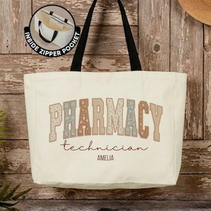 May include: A cream-colored canvas tote bag with black shoulder straps. The front features the word PHARMACY in a floral-patterned block font, with technician and AMELIA printed below. An inset circle shows an internal zipper pocket.
