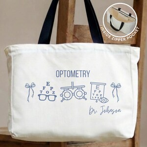 May include: A white canvas tote bag with black handles featuring blue graphics of optometry equipment, including an eye chart, phoropter, and glasses. The text reads OPTOMETRY and Dr. Johnson. An inset shows an inside zipper pocket.