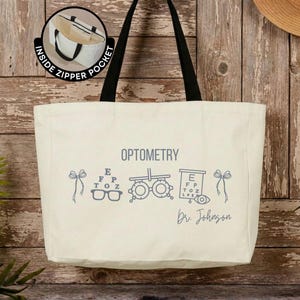 May include: A cream-colored canvas tote bag with black shoulder straps. The front features the text OPTOMETRY, an eye chart, a phoropter, and the name Dr. Johnson in blue. A circular inset shows an interior view labeled INSIDE ZIPPER POCKET.