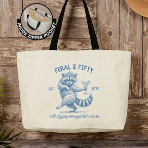 May include: A cream-colored canvas tote bag with black handles and an inside zipper pocket. The front features a blue graphic of a raccoon wearing a crown, holding a martini glass, with the text FERAL & FIFTY EST. 1976 still digging through life's trash.