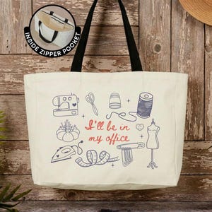 May include: A cream-colored canvas tote bag with black shoulder straps and an inside zipper pocket. The front features blue line art of sewing tools including a sewing machine, scissors, thimble, thread, iron, and dress form, with the red text I'll be in my office.