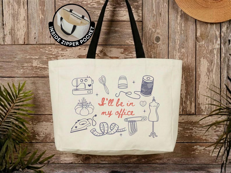 May include: A cream-colored canvas tote bag with black shoulder straps and an inside zipper pocket. The front features blue line art of sewing tools including a sewing machine, scissors, thimble, thread, iron, and dress form, with the red text I'll be in my office.