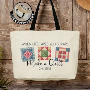 May include: A cream-colored canvas tote bag with black shoulder straps. The front features three colorful quilt block illustrations, the text "WHEN LIFE GIVES YOU SCRAPS Make a Quilt" and the name "CHRISTINE". Includes an inside zipper pocket.