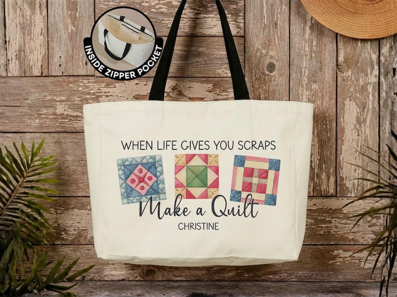 Puede incluir: Un bolso de lona color crema con correas negras para el hombro. El frente presenta tres ilustraciones de bloques de edred&oacute;n coloridos, el texto "WHEN LIFE GIVES YOU SCRAPS Make a Quilt" y el nombre "CHRISTINE". Incluye un bolsillo interior con cremallera.