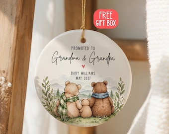 Può includere: Un ornamento rotondo in ceramica bianca appeso a un cordino dorato. Presenta un'illustrazione di tre orsi seduti in un campo con montagne sullo sfondo. Il testo recita: PROMOTED TO Grandma & Grandpa, BABY WILLIAMS, MAY 2027 e un cuore rosso.