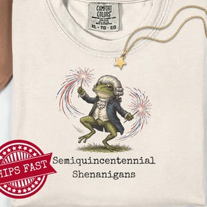 Könnte beinhalten: Cremefarbenes T-Shirt mit einer Cartoon-Froschfigur in einem Kolonialoutfit, die Wunderkerzen mit roten, weißen und blauen Feuerwerkskörpern hält. Der Text "Semiquincentennial Shenanigans" ist darunter gedruckt. Ein goldener Sternanhänger hängt an einer goldenen Kette.
