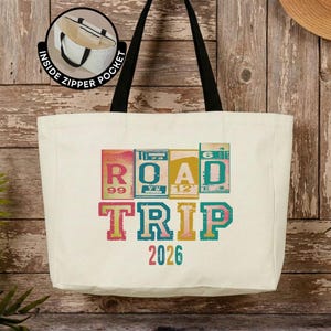 May include: A cream-colored canvas tote bag with black shoulder straps. The front features a graphic with the text ROAD TRIP 2026 in colorful, blocky, vintage-style lettering. A circular inset shows an interior view labeled INSIDE ZIPPER POCKET.