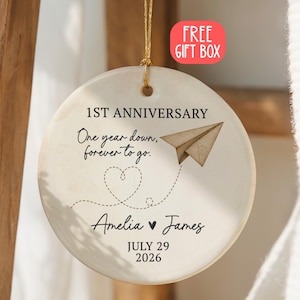 Puede incluir: Un adorno redondo de cerámica blanca colgado de un cordón dorado. Presenta el texto 1ST ANNIVERSARY, One year down, forever to go., Amelia, James y JULY 29 2026. Un gráfico de avión de papel marrón y una línea de corazón punteada están impresos.