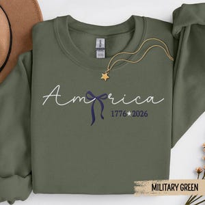 May include: Military green sweatshirt with the word "America" in script, a blue bow, and the numbers "1776 2026". A gold star pendant hangs from a gold chain. A brown hat is in the background.