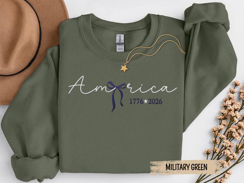 May include: Military green sweatshirt with the word "America" in script, a blue bow, and the numbers "1776 2026". A gold star pendant hangs from a gold chain. A brown hat is in the background.