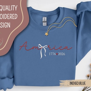 May include: Indigo blue sweatshirt with the word "America" embroidered in red script, a white bow, and the dates "1776-2026." A gold star necklace is draped across the front. The words "High Quality Embroidered Design" are at the top.