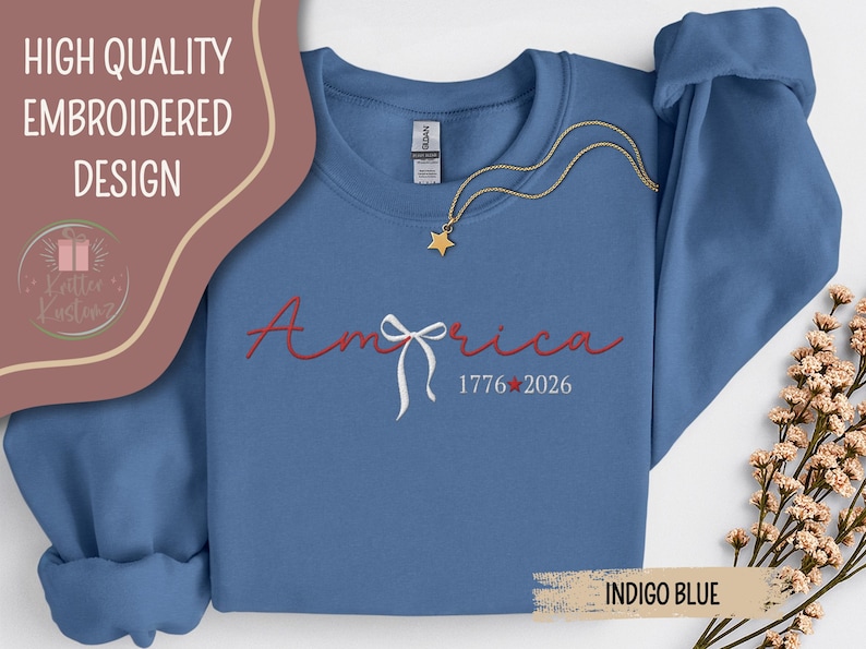 May include: Indigo blue sweatshirt with the word "America" embroidered in red script, a white bow, and the dates "1776-2026." A gold star necklace is draped across the front. The words "High Quality Embroidered Design" are at the top.