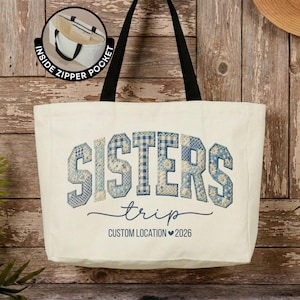 May include: A cream-colored canvas tote bag with black shoulder straps. The front features the word SISTERS in blue and white patchwork-style letters, with the word trip and the text CUSTOM LOCATION 2026 below. An inset shows an inside zipper pocket.