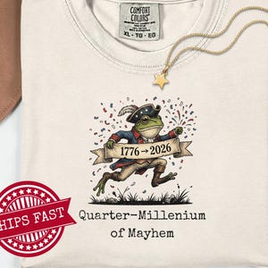 Könnte beinhalten: Cremefarbenes T-Shirt mit einer Cartoon-Froschfigur in einem Dreispitzhut und Kolonialkleidung, die ein Banner mit "1776 -> 2026" hält. Der Text "Quarter-Millennium of Mayhem" steht darunter. Eine goldene Sternkette ist sichtbar.