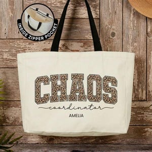May include: A cream-colored canvas tote bag with black shoulder straps. The front features the word CHAOS in a leopard print pattern, the word coordinator in black script, and the name AMELIA in black text. An inset shows an inside zipper pocket.