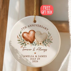 May include: A round white ceramic ornament hanging by a gold string. The design features two hearts, one brown and one cream-colored, surrounded by floral greenery. Text reads 7TH ANNIVERSARY, Forever & Always, AMELIA & JAMES, JULY 29 2026.