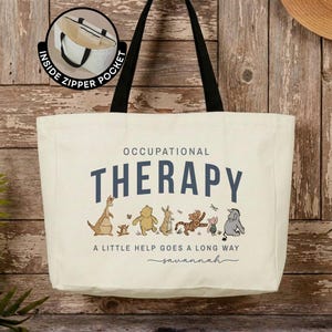 May include: A cream-colored canvas tote bag with black shoulder straps. The front features the text OCCUPATIONAL THERAPY, A LITTLE HELP GOES A LONG WAY, and the name Savannah, with illustrations of Winnie the Pooh characters.