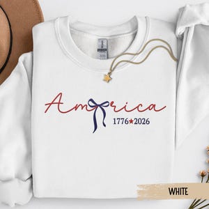 May include: White crewneck sweatshirt with the word "America" in red script, a blue bow, and the dates "1776*2026". A gold necklace with a star pendant is draped on the sweatshirt.