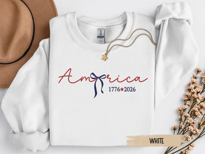 May include: White crewneck sweatshirt with the word "America" in red script, a blue bow, and the dates "1776*2026". A gold necklace with a star pendant is draped on the sweatshirt.