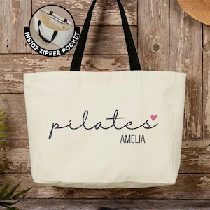 Pu&ograve; includere: Una borsa tote in tela color crema con spallacci neri. La parte anteriore presenta la parola pilates in corsivo nero, un piccolo cuore rosa e il nome AMELIA in lettere maiuscole nere. Un riquadro mostra una tasca interna con cerniera.