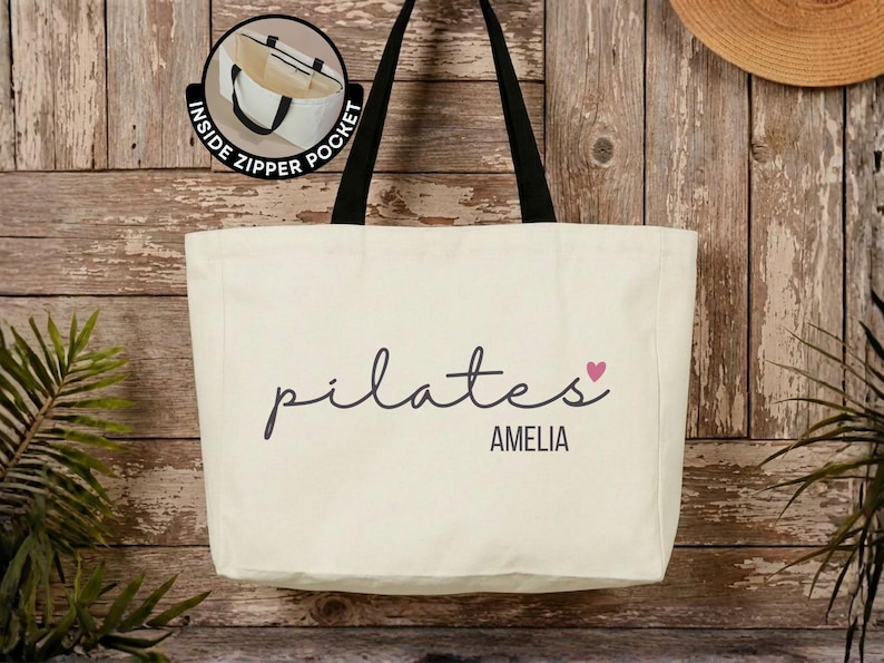 Pu&ograve; includere: Una borsa tote in tela color crema con spallacci neri. La parte anteriore presenta la parola pilates in corsivo nero, un piccolo cuore rosa e il nome AMELIA in lettere maiuscole nere. Un riquadro mostra una tasca interna con cerniera.