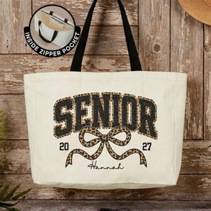 May include: A cream-colored canvas tote bag with black handles. The front features the word SENIOR in black leopard print, a leopard print bow, the numbers 20 and 27, and the name Hannah. An inset shows an inside zipper pocket.