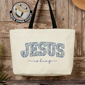 May include: A cream-colored canvas tote bag with black shoulder straps. The front features the text JESUS in a blue floral pattern, with the words is king written below in a blue script font. A circular inset shows an inside zipper pocket.