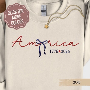 May include: Cream-colored sweatshirt with the word "America" in red script, a blue bow, and the numbers "1776*2026" in blue. A gold star pendant hangs from a gold chain. A tag reads "SAND".