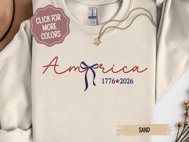 May include: Cream-colored sweatshirt with the word "America" in red script, a blue bow, and the numbers "1776*2026" in blue. A gold star pendant hangs from a gold chain. A tag reads "SAND".
