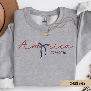 May include: Sport grey sweatshirt with the word "America" in red script, a blue bow, and the numbers "1776*2026" in navy blue. A gold star pendant necklace rests on the sweatshirt.