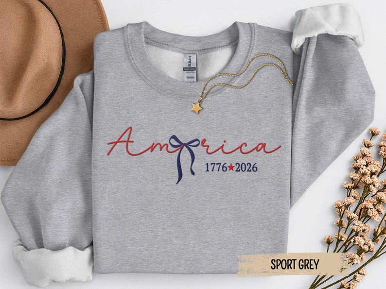 May include: Sport grey sweatshirt with the word "America" in red script, a blue bow, and the numbers "1776*2026" in navy blue. A gold star pendant necklace rests on the sweatshirt.