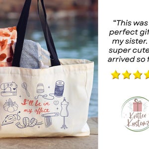 May include: A cream-colored canvas tote bag with black handles, featuring blue and red line art of sewing tools and the text "I'll be in my office." The bag contains a folded orange patterned fabric and a blue and white geometric patterned textile.